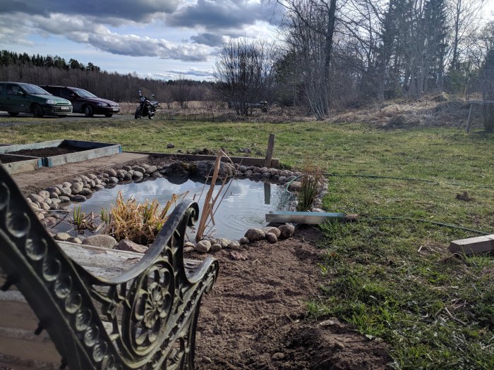 Nyflyttad trädgårdsdamm med stenar och växter, promenadplattor och pallkragar med jord, Honda motorcykel i bakgrunden.