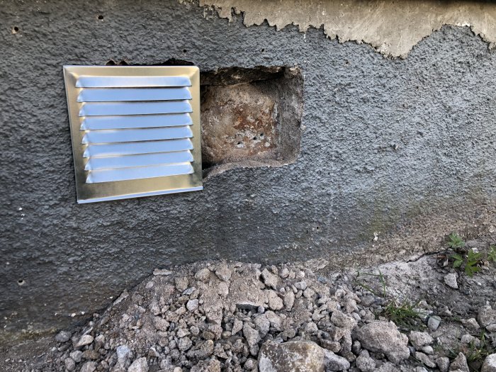 Nyinstallerat ventilgaller på en skadad grundmur med ett ofärdigt hål och lösa stenar vid foten av muren.