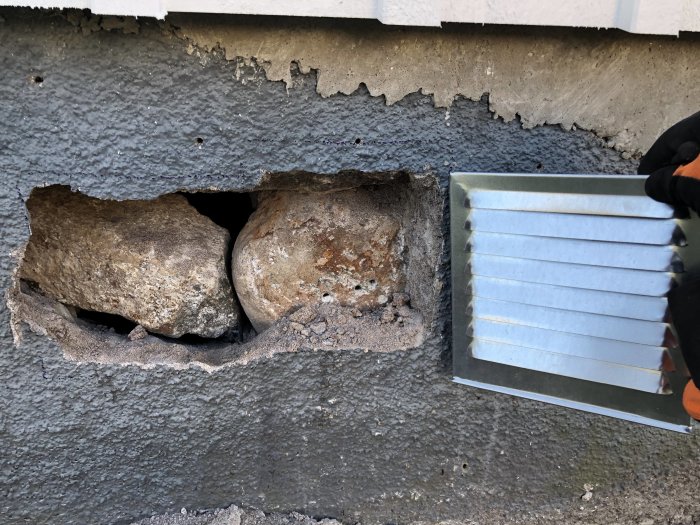 Person som håller i ett ventilgaller bredvid ett bilat hål i en grundmur av natursten och sparsamt cement från 1929.