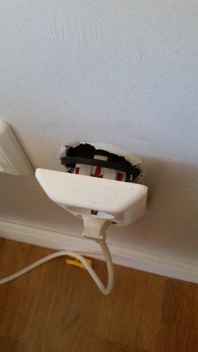 Sprucken installationsdosa bakom ett öppet vägguttag med ansluten sladd.