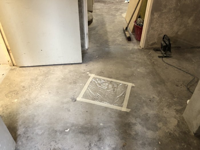 Fukttest på betonggolv med en liten kvadrat av plastfolie tejpfast mot golvet i ett delvis renoverat rum.