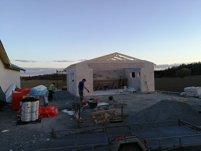 Obyggt garage med öppen takkonstruktion och byggmaterial spridda på marken, två personer jobbar med projektet.