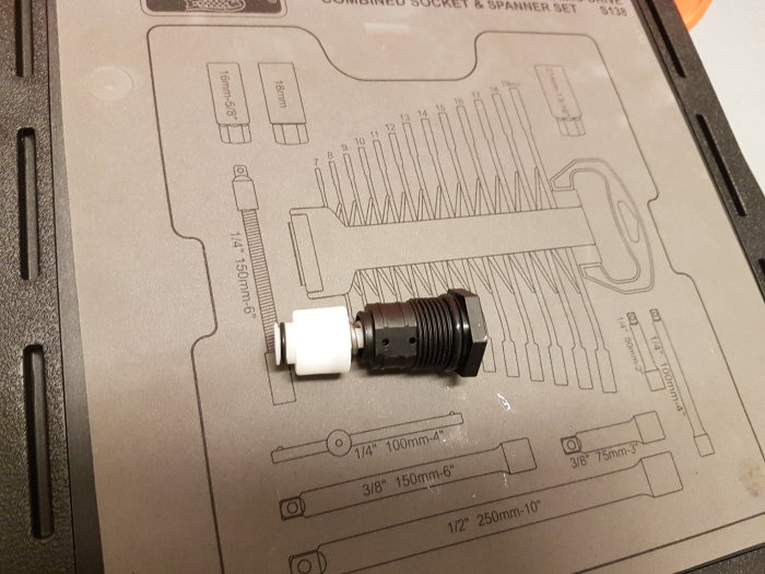 Svart cylindrisk komponent med vit kolv på en verktygslåda, möjligt byttdel för tvättmaskinsreparation.