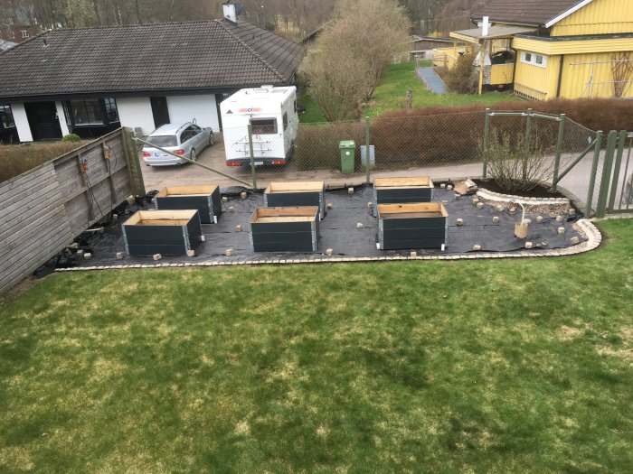 Odlingslådor på svart fiberduk med omgivande stenar, med staket och hus i bakgrunden, nygrävd mark synlig.