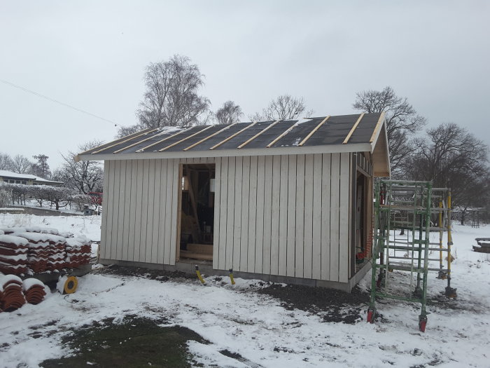 Halvfärdigt trädgårdsskjul med uppsatt panel och takstolar täckta med takpapp i snöigt landskap.