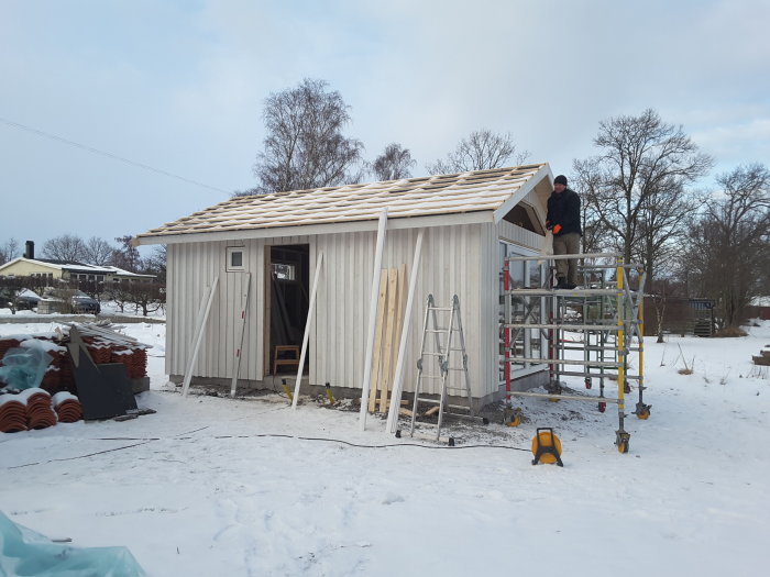 Byggarbetsplats med ofärdigt trähus och person på byggställning i snöig miljö.