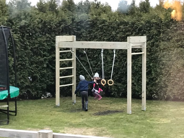 Två barn leker på en gungställning med ringar i en trädgård omgiven av häck.