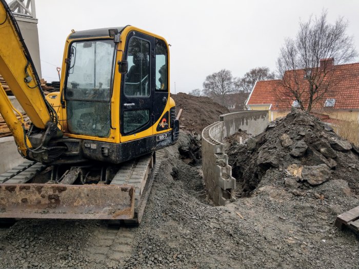 Gul grävmaskin nära en delvis byggd böjd stödmur och högar av grus och jord.