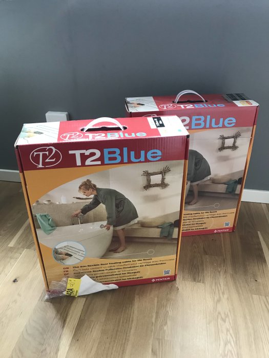 Två förpackningar av golvvärmekabel T2Blue på ett trägolv mot en grå vägg.