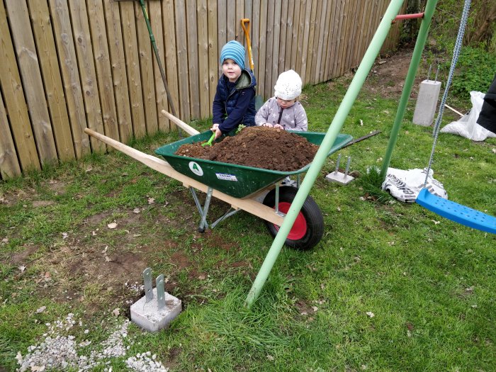 Två barn leker i en skottkärra full med jord bredvid betongplintar och en gungställning.