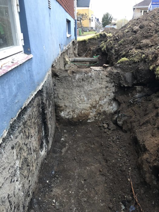Grävd grund vid hus med olika grunder, synligt rör och tegelvägg, under pågående dräneringsarbete.