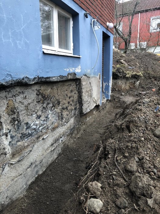 Grävd grund vid en blå byggnad med synlig äldre och nyare del av fundamentet samt ett grävt dike.