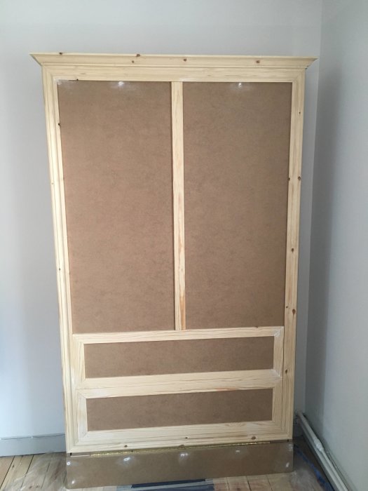 Obehandlat trä-sängskåp i ett rum, monterat mot en vägg, ej fullständigt färgat och utan handtag.