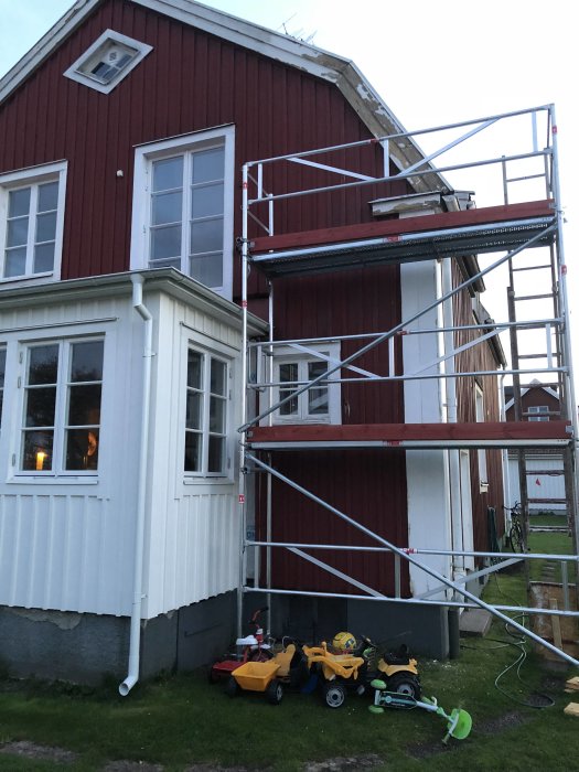 Ställning monterad intill en röd träfasad på ett hus för renoveringsarbete, barnleksaker syns i förgrunden.