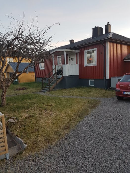 Ett rött trähus med vita knutar och en källartrappa, gräsmatta med liten nivåskillnad för tänkt utbyggnad.