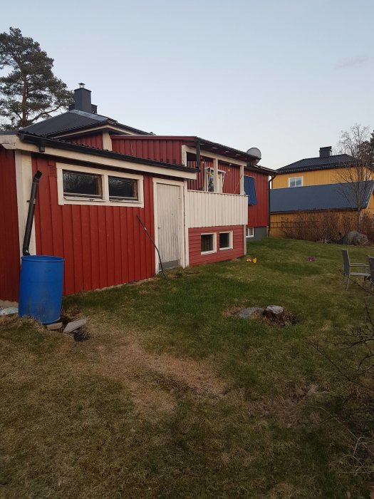 Rödmålat trähus med en källare, omgiven av gräsmatta med liten nivåskillnad i bakgården.