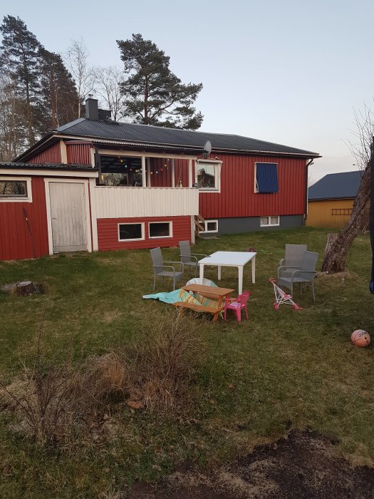 Trädgård med gräsmatta och nivåskillnader bakom en röd trävilla, möbelgrupp och barnleksaker synliga.