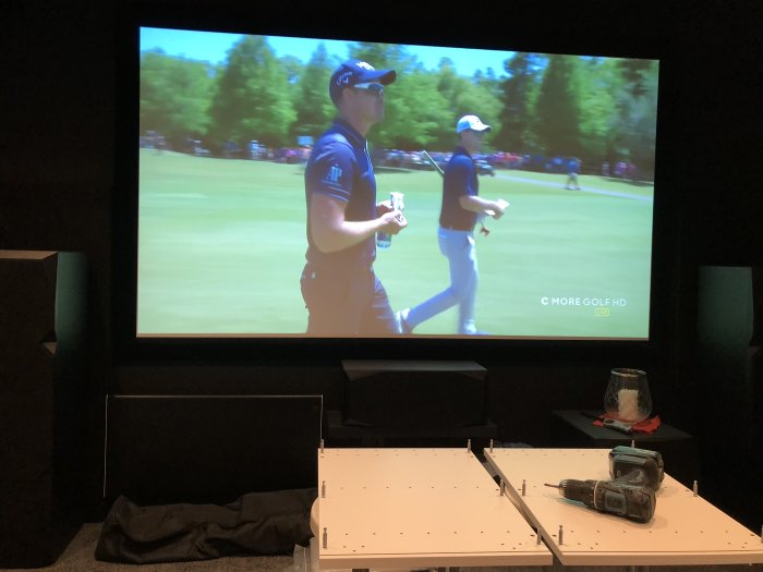 Överskåp under konstruktion på golv med skruvdragare och verktyg framför en TV som visar golf.