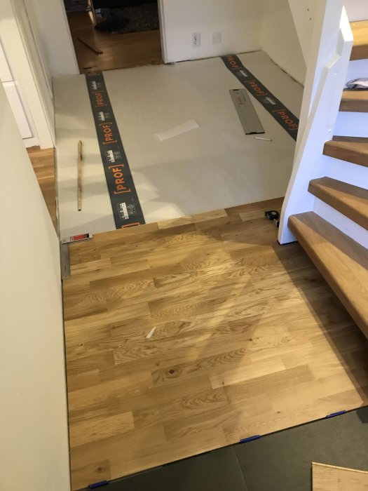 Nytt trägolv under installation i en hall med en bit anpassad runt trappan, underlägg synligt och verktyg på golvet.