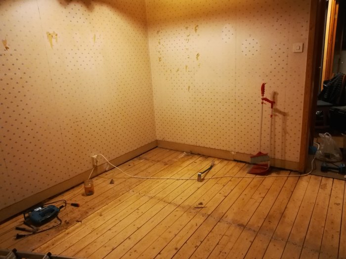 Rum under renovering med blottat, massivt trägolv och väggar med gammal tapet. Verktyg och renoveringsmaterial synliga.