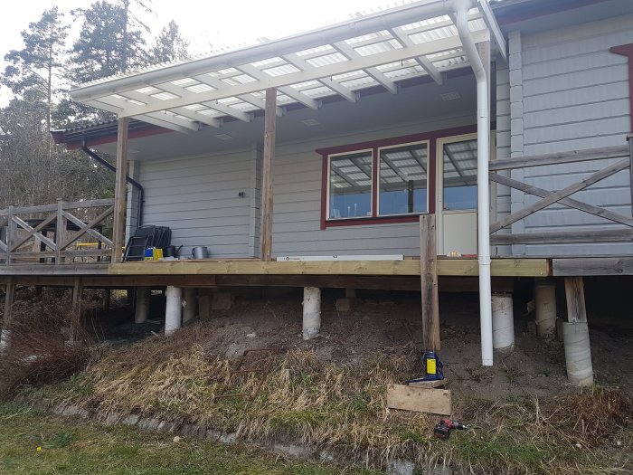 Altan under ombyggnation till uterum med vitmålade plintar och delvis lagt golv, del av hus och trädgård syns.