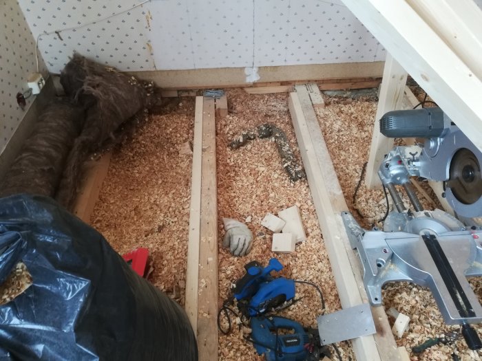 Renoveringsarbete på golv med kutterspån, isolering och träreglar synliga, samt verktyg utspridda.