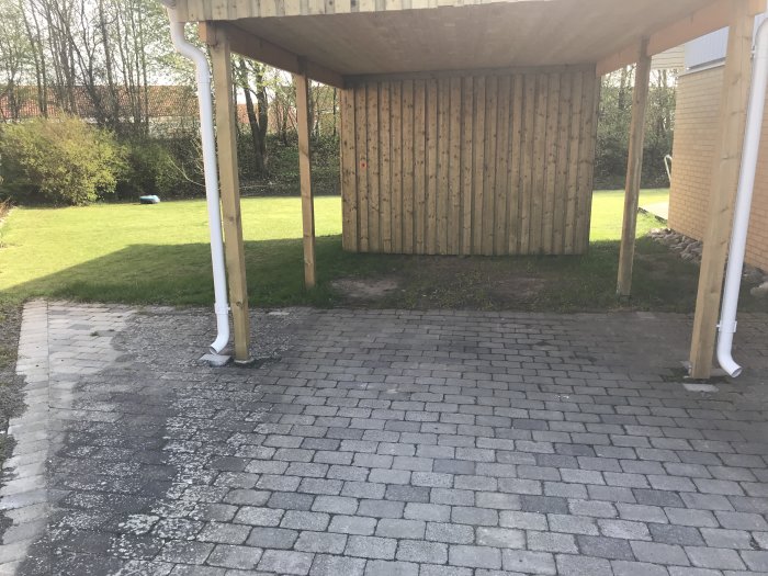 Uppfart med grå "byasten" under carport, angränsande gräsplätt och förråd i bakgrunden.