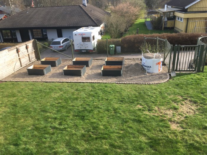Nytt grus i en stor säck i en trädgård med upplagda odlingslådor på grusgång, med hus och husbil i bakgrunden.