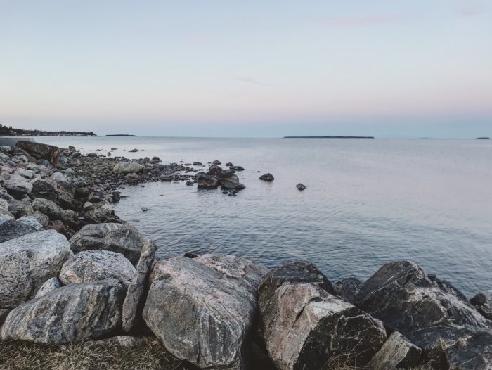 Kvällsutsikt över en lugn kustlinje med stora stenar i förgrunden och en blek himmel ovanför.