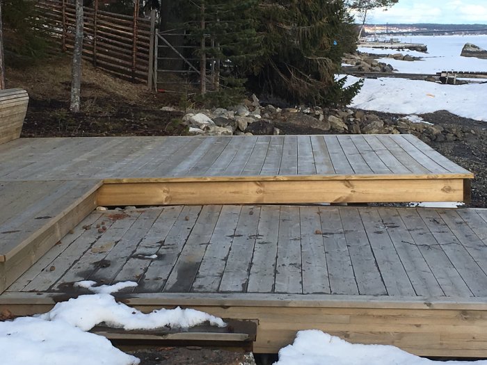 Träbrygga med sättningar omgiven av snö och terräng med vatten i bakgrunden.