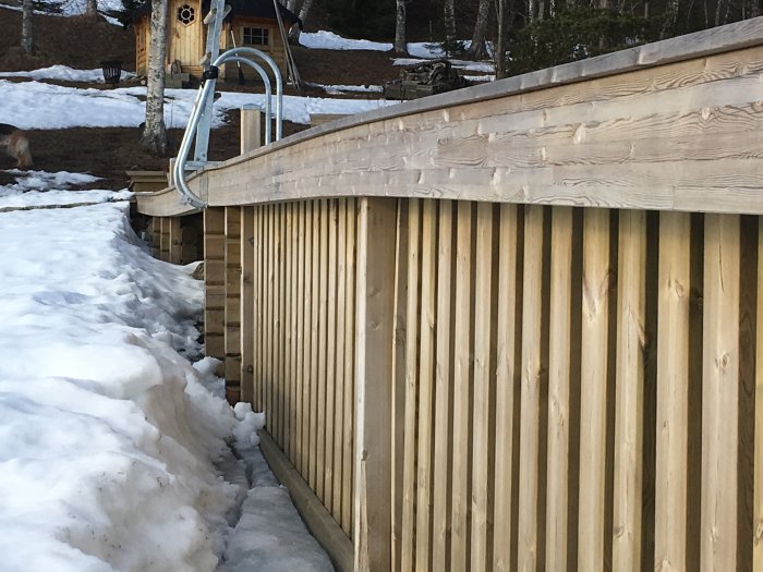 Träbrygga med sättningsskador nära en snödriva, med omgivande smältande snö och ett hus i bakgrunden.