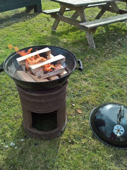 Nytänd grill med brinnande ved på en gräsmatta, bredvid en picknickbänk och avtaget grilllock.