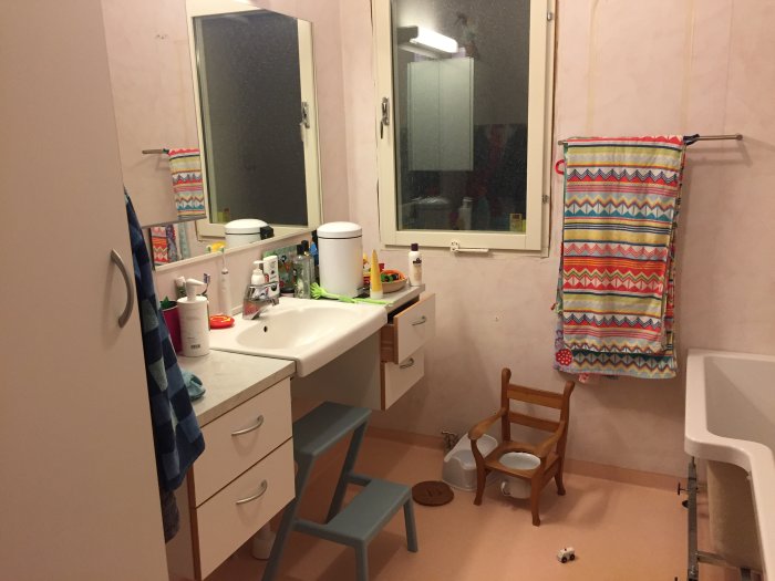 Gammalt badrum före renovering med rosa väggar, handfat, spegelskåp och våtrumsmatta på golvet.