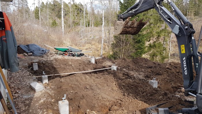 Grävmaskin vid arbete med plintfundament och grovarbeten för växthus på en tomt.