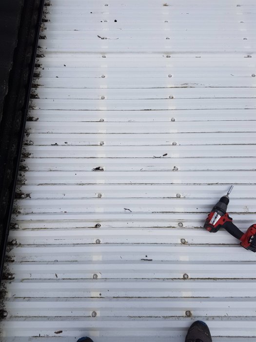 Vitt tak med tätt fördelade skruvar och en röd borrmaskin ligger på trallen, fotspetsar skymtar nederst.