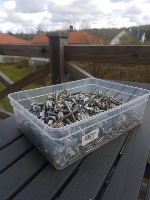 Genomskinlig låda med många urdragna skruvar på ett utomhusträdäck, trädgård i bakgrunden.