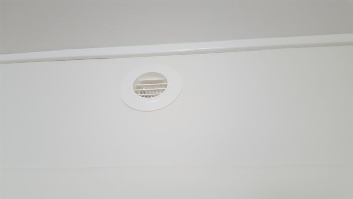 Rund ventil i vit vägg ovanför en list, med synligt ventilationsfilter inuti.