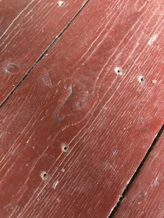 Slitet rödfärgat trägolv med tydliga ådringsmönster och små spikhål, tecken på generationers användning.