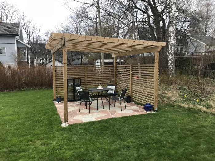 Träpergola med plattlagd uteplats och sittgrupp på stensättning som ligger ovanför en gräsmatta.