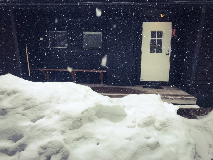 Täckt med snö, en husfasad med en vit dörr och två fönster med snöflingor som faller tätt i luften.