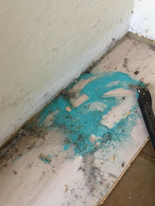 Utspridda turkosfärgade byggspån och damm i ett hörn på ett trägolv bredvid en vit vägg.
