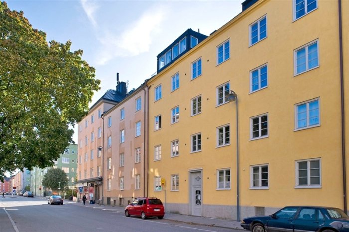 Gult flerbostadshus från 1930-talet på en stadsgata med bilar parkerade utanför och träd i bakgrunden.