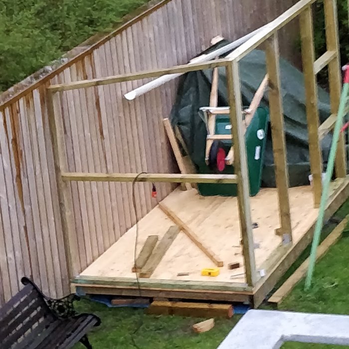 Pågående bygge av ett ved- och redskapsskjul med lutande regelverk framför ett befintligt plank.