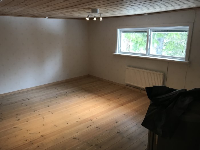 Tomt rum med trägolv, tapetserade väggar, takbjälkar och ett fönster med utsikt över träd.