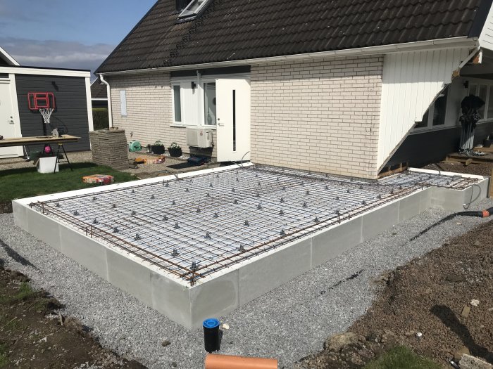 Grundplatta med installerad armering och golvvärme bredvid ett hus, med kabelrör som sträcker sig mot en liten byggnad.