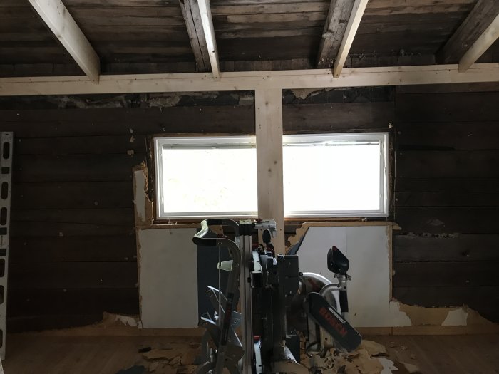Renovering av rum med träväggar och synliga takbjälkar, fönster och arbetsutrustning på golvet.