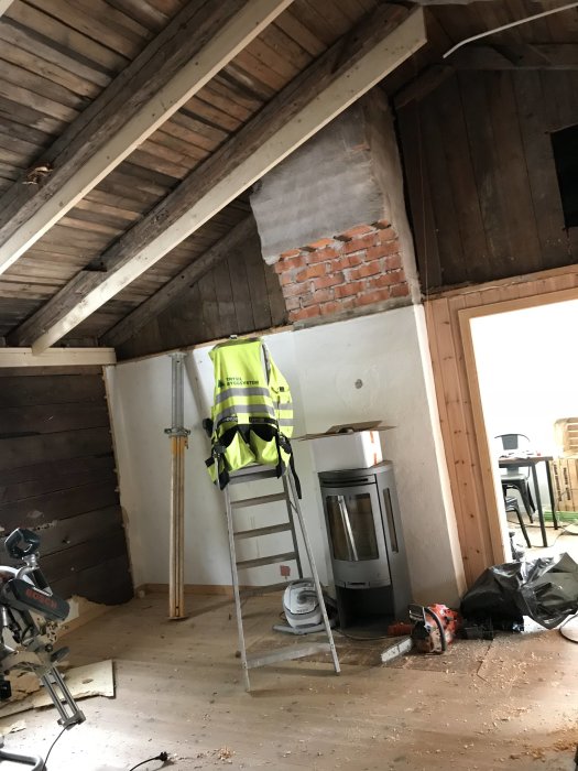 Renoveringsarbete i ett rum med synliga takbjälkar och en ofärdig tegelskorsten, verktyg och byggmaterial syns.