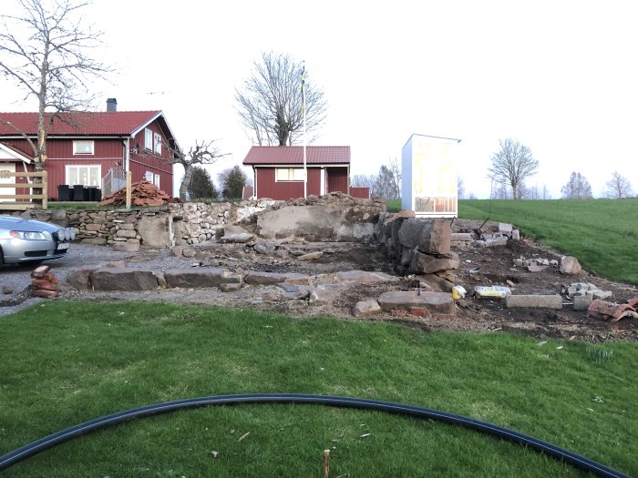 Röd stuga bakom en oordnad byggarbetsplats med stenmur och röjning, dagen före byggstart.