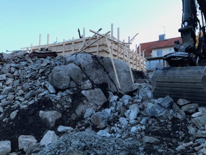 Gjutform för stödmur under uppbyggnad bland stora stenar och grus med grävmaskin i förgrunden.