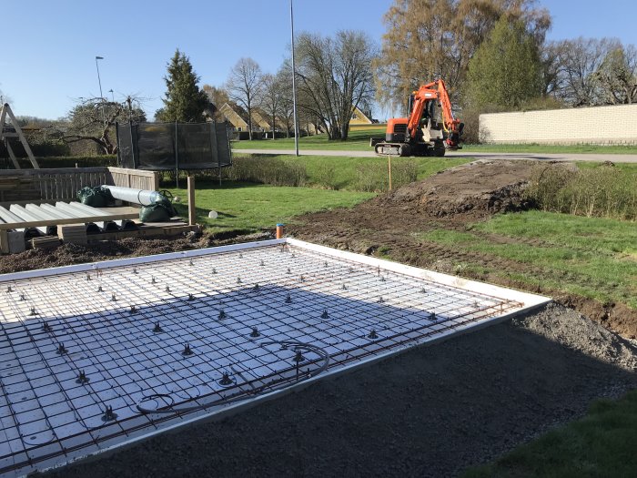 Grävmaskin fyller på material runt en ojuten grund med armeringsnät, i ett soligt bostadsområde.